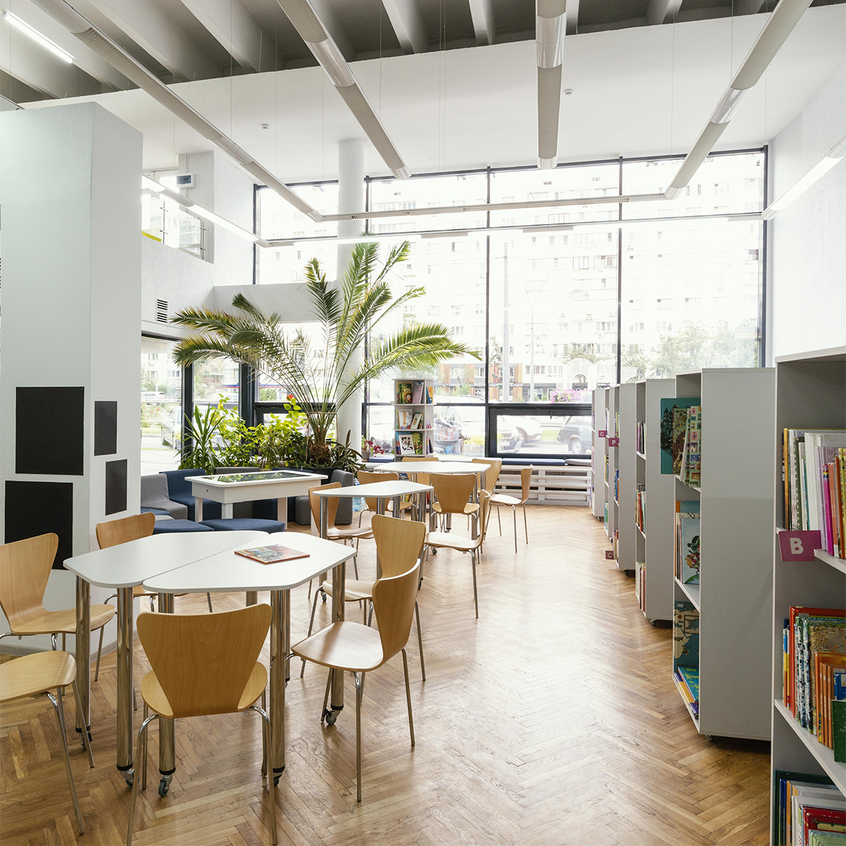 Heller, moderner Bildungsraum mit Tischen, Holzstühlen und offenen Bücherregalen vor großer Fensterfront